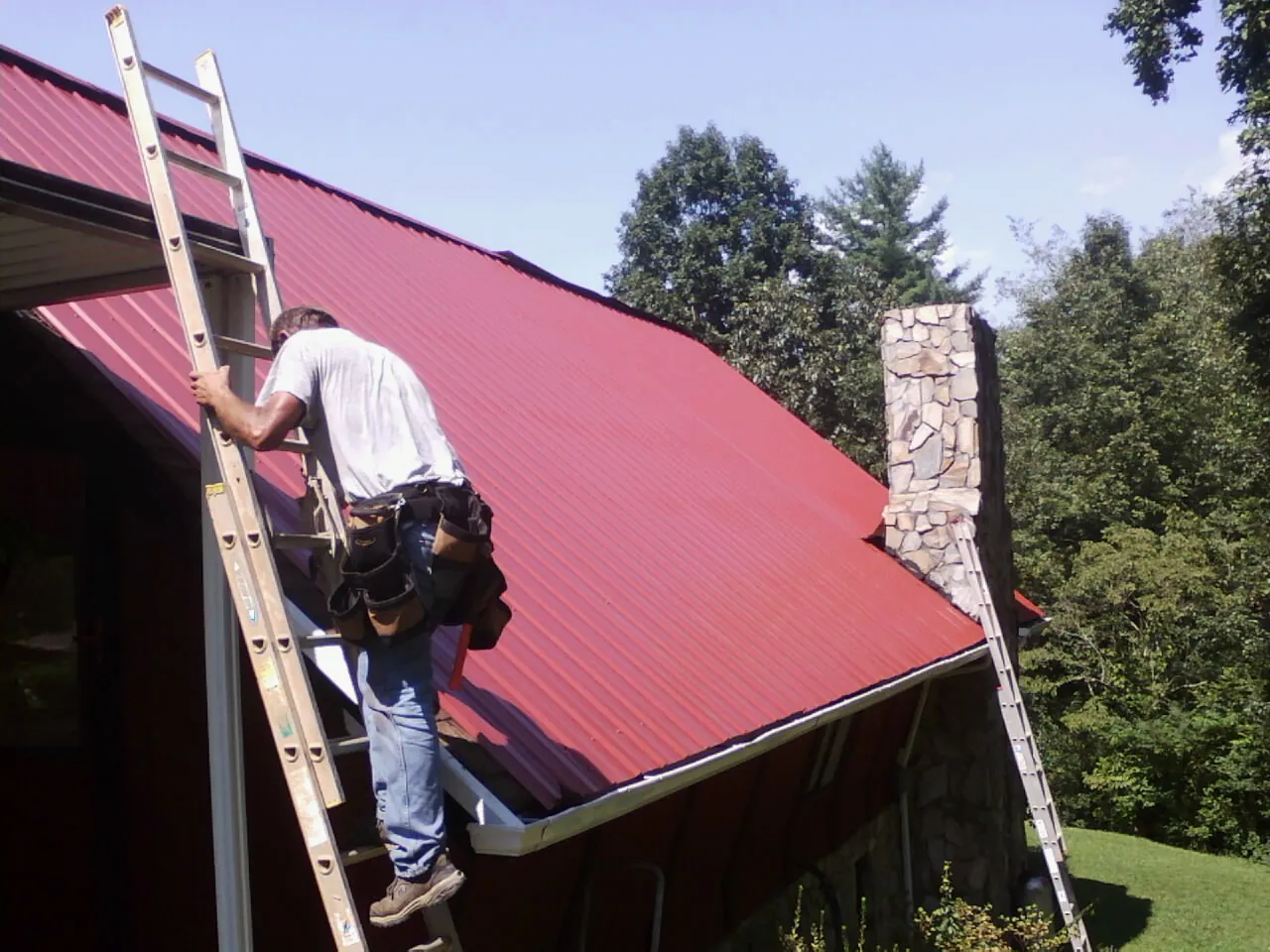 Roofer on a ladder beside a red metal roof and stone chimney — Soffit & Fascia Repair by Roofing Brothers in Swainsboro